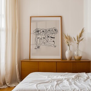 Puede incluir: Una impresi&oacute;n art&iacute;stica enmarcada que presenta un dibujo lineal de tres figuras con cabezas florales. La obra de arte es en blanco y negro, sobre un fondo beige, y est&aacute; enmarcada en un marco de madera marr&oacute;n claro. La impresi&oacute;n se muestra encima de un mueble de madera.
