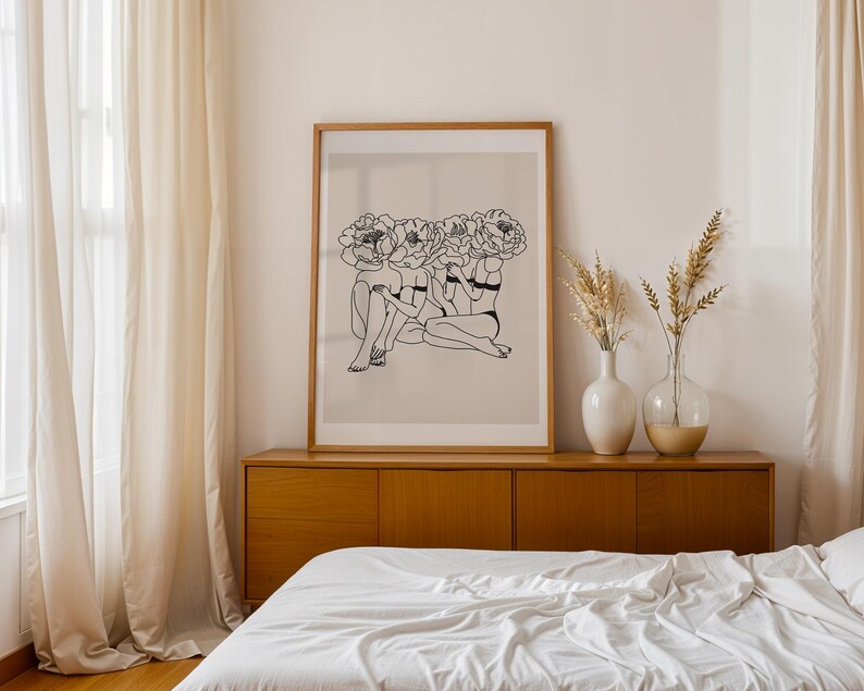 Puede incluir: Una impresi&oacute;n art&iacute;stica enmarcada que presenta un dibujo lineal de tres figuras con cabezas florales. La obra de arte es en blanco y negro, sobre un fondo beige, y est&aacute; enmarcada en un marco de madera marr&oacute;n claro. La impresi&oacute;n se muestra encima de un mueble de madera.