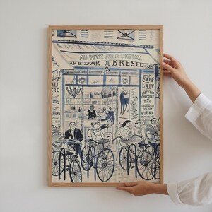 May include: A framed art print featuring a blue and white ink illustration of a Parisian cafe scene titled AU PETIT FER A CHEVAL CAFE BAR DU BRESIL. The artwork depicts people sitting at tables with bicycles parked in front of the storefront.