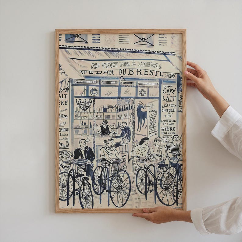 May include: A framed art print featuring a blue and white ink illustration of a Parisian cafe scene titled AU PETIT FER A CHEVAL CAFE BAR DU BRESIL. The artwork depicts people sitting at tables with bicycles parked in front of the storefront.