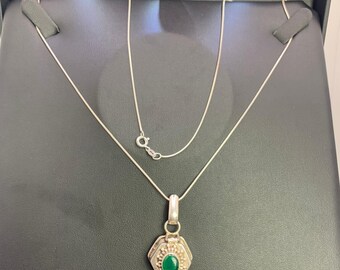 May include: A silver necklace with a hexagonal pendant featuring a central oval green stone. The pendant is set in a decorative silver frame. The necklace is displayed on a black surface.