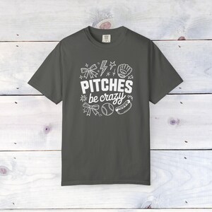 Op de afbeelding: Donkergrijs t-shirt met de witte tekst "Pitches be crazy" en honkbal-thema afbeeldingen, waaronder een honkbalhandschoen, honkbal en hotdog. Het shirt heeft een ronde hals en korte mouwen.