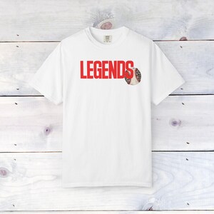 Op de afbeelding: Wit T-shirt met het woord "LEGENDS" in rode blokletters. Een honkbal met een rode naad en een patroon staat naast het woord "LEGENDS". Het shirt is gemaakt van een zacht materiaal.