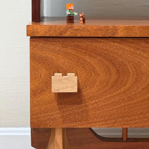 May include: A wooden cabinet with a drawer featuring a unique, block-shaped wooden handle. The cabinet has a light brown finish and sits on four legs.