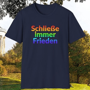 May include: A navy blue t-shirt with the German phrase "Schließe immer Frieden" in rainbow colors. The text translates to "Always make peace". The shirt is displayed outdoors with a scenic background.