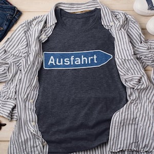 May include: A dark blue t-shirt with a white graphic of a blue and white German road sign that says "Ausfahrt" which means "Exit" in English.