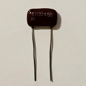 May include: A dark brown rectangular capacitor with two silver leads. The capacitor has the text "5100 ±5%" and the letter "M" printed on it. This electronic component is used in circuits.