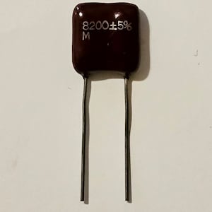 May include: A dark brown, rectangular electronic component with two silver leads. The component has the text "8200±5%" and "M" printed on it. The component is likely used in electronics.