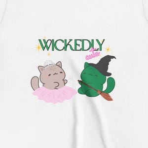 May include: White t-shirt featuring a whimsical design with two cartoon cats. One cat wears a tutu and crown, the other a witch hat and holds a broom. The text "WICKEDLY cute" is above the cats.