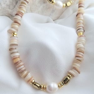 Könnte beinhalten: Eine weiße und beige Muschelkette mit goldenen Akzenten und einer großen weißen Perle. Die Kette liegt auf einem weißen Stoffhintergrund.
