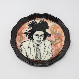May include: Round ceramic plate with a black scalloped edge and a central image of a person in a suit with a tie. The background is peach with gold dollar signs. The plate has a black and orange striped border.