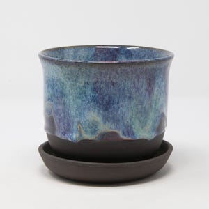 May include: A ceramic planter with a smooth, dark brown base and a glossy, dripped glaze finish in shades of blue and purple. The cylindrical pot sits securely on a matching dark brown circular saucer, creating a two-piece set.