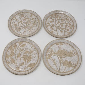 May include: Four round ceramic coasters with floral designs. Each coaster features a different botanical pattern in a light brown color against a speckled white background. The designs include leaves, flowers, and other plant elements.