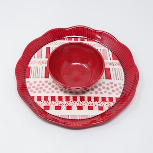 May include: A red ceramic serving set featuring a round tray with a scalloped edge and a small bowl. The tray has a red and white patterned design with pink accents. The bowl is solid red.