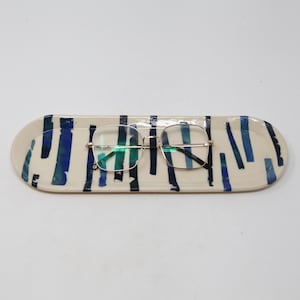 May include: A rectangular ceramic tray with a cream-colored base and blue brushstroke design. A pair of eyeglasses with a gold-colored frame rests on the tray. The tray is approximately 10 inches long.