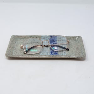 May include: A pair of eyeglasses with a gold-colored frame rests on a rectangular ceramic tray. The tray has a speckled gray surface with a blue and white floral pattern in the center.