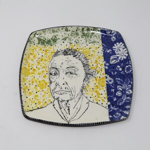 May include: A square ceramic plate with a black and white line drawing of a woman's face. The background is a yellow and green floral pattern with black polka dots. The right side of the plate has a blue and white floral pattern.