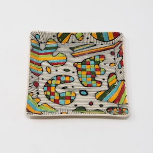 May include: A square ceramic tray with a white background and a colorful abstract design. The design features geometric shapes, stripes, and dots in shades of red, yellow, green, blue, and black.