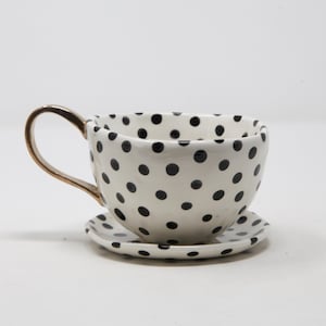 May include: A white ceramic teacup and saucer set with black polka dots. The teacup has a gold-colored handle. The saucer is round and also features black polka dots. The set is designed for serving tea or coffee.