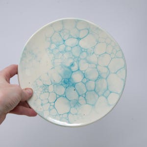 May include: A round, off-white ceramic plate with a blue bubble pattern. The plate has a glossy finish and is held by a hand. The blue pattern is concentrated in the center and fades towards the edges.