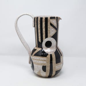May include: A ceramic pitcher with a white handle and a speckled, geometric design in black and tan. The pitcher features a circular accent and a spout for pouring.