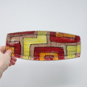 May include: Rectangular decorative tray with a colorful geometric design. The tray features a pattern of red, yellow, and orange rectangles and lines, outlined with black dots. The tray is held by a hand against a white background.