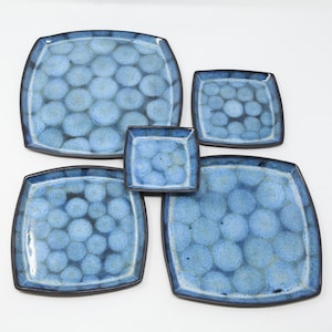 May include: A set of five square ceramic plates in varying sizes. The plates are a mottled blue with a pattern of overlapping circles and a dark brown rim. The plates are arranged on a white background.