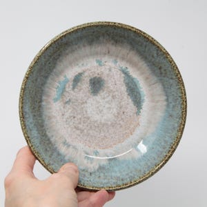 May include: A round ceramic bowl featuring a reactive glaze with a blend of light blue, white, and soft pink tones. The rim is textured with a speckled brown finish, and the interior surface displays a smooth, glossy gradient transition between the colors.