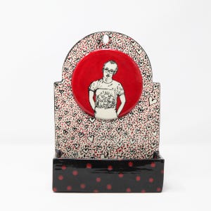 May include: A ceramic wall mounted shelf with a red circle containing a black and white illustration of a person wearing glasses and a t-shirt with a graphic. The background is white with black and red hearts.