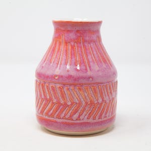 May include: A ceramic vase with a glossy pink and orange glaze. The upper section features vertical carved lines, while the lower section displays a horizontal band of repeating diagonal chevron patterns, revealing the orange clay beneath the glaze.