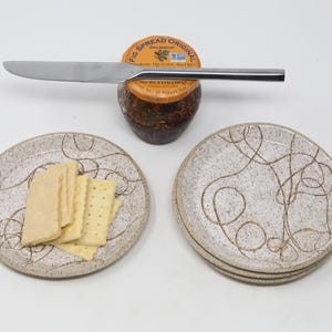 May include: A fig spread jar with a stainless steel knife resting on top. Two small ceramic plates with a cream-colored abstract design are in the foreground, one with crackers and cheese. The jar has a label that reads "Fig Spread Original".