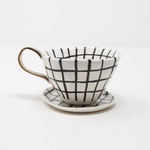 May include: White ceramic teacup and saucer set with a black grid pattern. The teacup has a gold-colored handle. The saucer is round and matches the teacup's design. The set is on a white background.