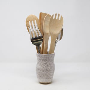 May include: A collection of kitchen utensils, including wooden spoons, a spatula, and a metal fork, are displayed in a speckled ceramic holder. The utensils are made of wood and stainless steel, and the holder is a light gray color.