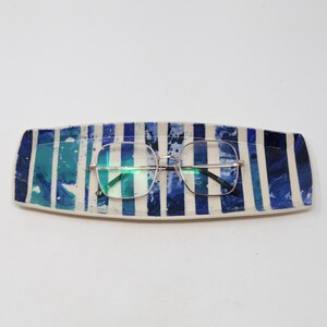May include: A rectangular ceramic eyeglasses holder with a blue and white striped pattern. The holder has a pair of square-framed eyeglasses resting on it. The color scheme includes shades of blue and white.
