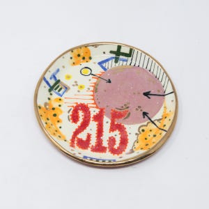 May include: A small white ceramic dish with a gold rim. The dish has a pink circle with black arrows pointing towards it. The number 215 is written in red on the dish. The dish is decorated with yellow, orange, and green paint.