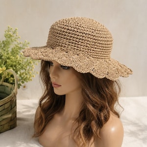 May include: A tan straw hat with a rounded crown and a wide, scalloped brim featuring a detailed crochet pattern. The hat is constructed from woven natural fibers, showcasing a textured surface and a decorative, fan-like edge design.