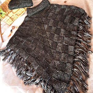 May include: A dark metallic gray knitted poncho featuring a basketweave pattern and long fringe along the edges, paired with a matching dark metallic gray knit beanie hat topped with a black faux fur pom-pom, both showcasing a shimmering texture.