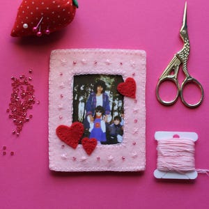 Puede incluir: Un marco de fotos de fieltro rosa con una foto familiar, adornos de corazón rojo y costuras decorativas. Un cojín de fresa rojo, tijeras doradas, hilo rosa y cuentas dispersas sobre un fondo rosa.