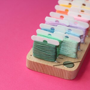 K&ouml;nnte beinhalten: Ein h&ouml;lzerner Faden-Organizer mit einem Nadelhalter. Der Organizer hat mehrere F&auml;cher zum Aufbewahren von verschiedenen Farben von Stickgarn. Das Garn ist in einem Regenbogenmuster angeordnet, beginnend mit Rot und endend mit Gr&uuml;n.