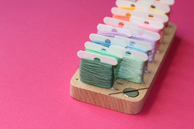 K&ouml;nnte beinhalten: Ein h&ouml;lzerner Faden-Organizer mit einem Nadelhalter. Der Organizer hat mehrere F&auml;cher zum Aufbewahren von verschiedenen Farben von Stickgarn. Das Garn ist in einem Regenbogenmuster angeordnet, beginnend mit Rot und endend mit Gr&uuml;n.