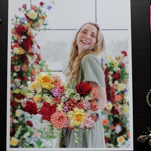 K&ouml;nnte beinhalten: Eine Frau mit langen blonden Haaren h&auml;lt einen Strau&szlig; bunter Seidenblumen. Die Blumen sind in verschiedenen Formen und Gr&ouml;&szlig;en angeordnet, darunter Rosen, G&auml;nsebl&uuml;mchen und Lilien. Die Frau tr&auml;gt ein hellgr&uuml;nes Kleid und l&auml;chelt.