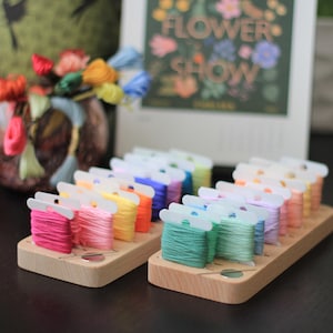 K&ouml;nnte beinhalten: Ein Holzorganizer mit Reihen von bunten Stickgarnrollen. Der Organizer hat einen durchsichtigen Plastikdeckel und ist mit "Flower Show" beschriftet.