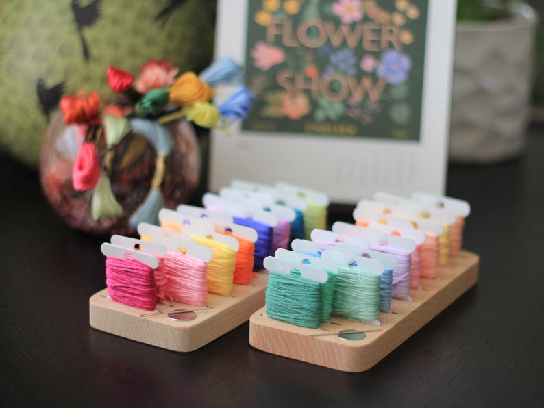 K&ouml;nnte beinhalten: Ein Holzorganizer mit Reihen von bunten Stickgarnrollen. Der Organizer hat einen durchsichtigen Plastikdeckel und ist mit "Flower Show" beschriftet.