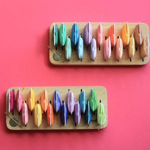 K&ouml;nnte beinhalten: Zwei Holzorganisatoren mit Reihen von bunten Garnrollen. Die Rollen sind in einem Regenbogenmuster angeordnet.