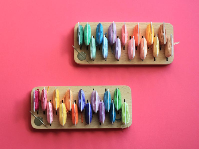 K&ouml;nnte beinhalten: Zwei Holzorganisatoren mit Reihen von bunten Garnrollen. Die Rollen sind in einem Regenbogenmuster angeordnet.