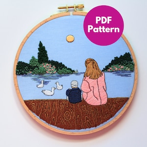 May include: Embroidered hoop art depicting a mother and child sitting on a wooden jetty overlooking a lake with ducks and a small island with trees. The sky is blue with a yellow sun. The text "PDF Pattern" is on a pink circle.