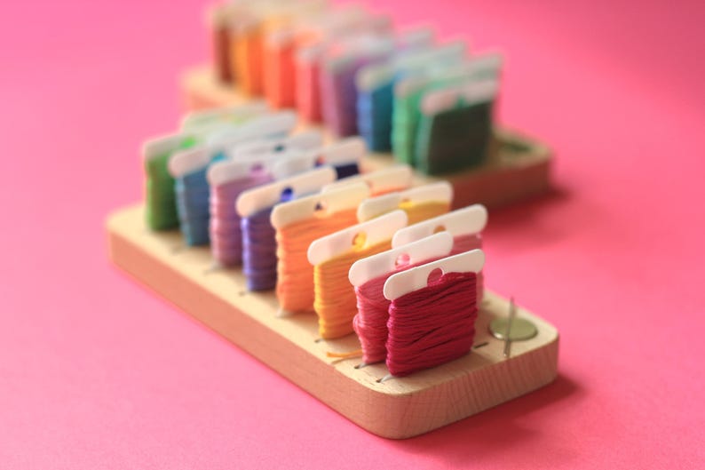 K&ouml;nnte beinhalten: Ein Holzorganizer mit Reihen von bunten Stickgarnrollen. Die Rollen sind in einem Regenbogenmuster angeordnet und werden durch transparente Plastikclips gehalten.