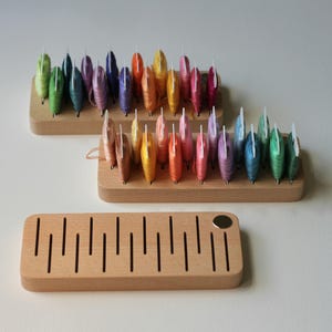 K&ouml;nnte beinhalten: Holzaufbewahrung f&uuml;r Stickgarn, mit regenbogenfarbenen Garnrollen in jedem Organizer. Die Organizer sind aus hellem Holz und haben eine nat&uuml;rliche Oberfl&auml;che.