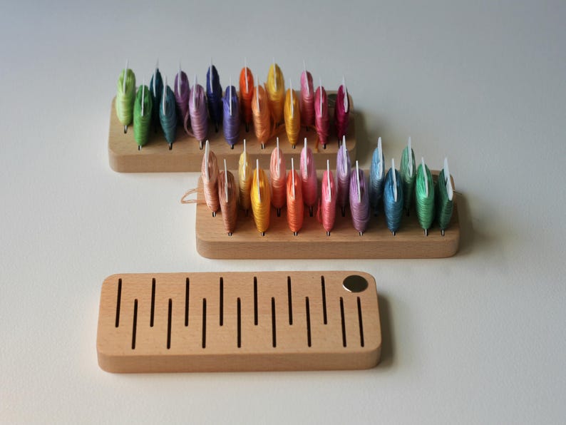 K&ouml;nnte beinhalten: Holzaufbewahrung f&uuml;r Stickgarn, mit regenbogenfarbenen Garnrollen in jedem Organizer. Die Organizer sind aus hellem Holz und haben eine nat&uuml;rliche Oberfl&auml;che.