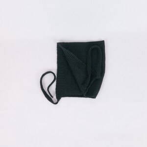 May include: Black knitted baby bonnet with a tie string. The bonnet is folded in half and the tie string is looped around the top.